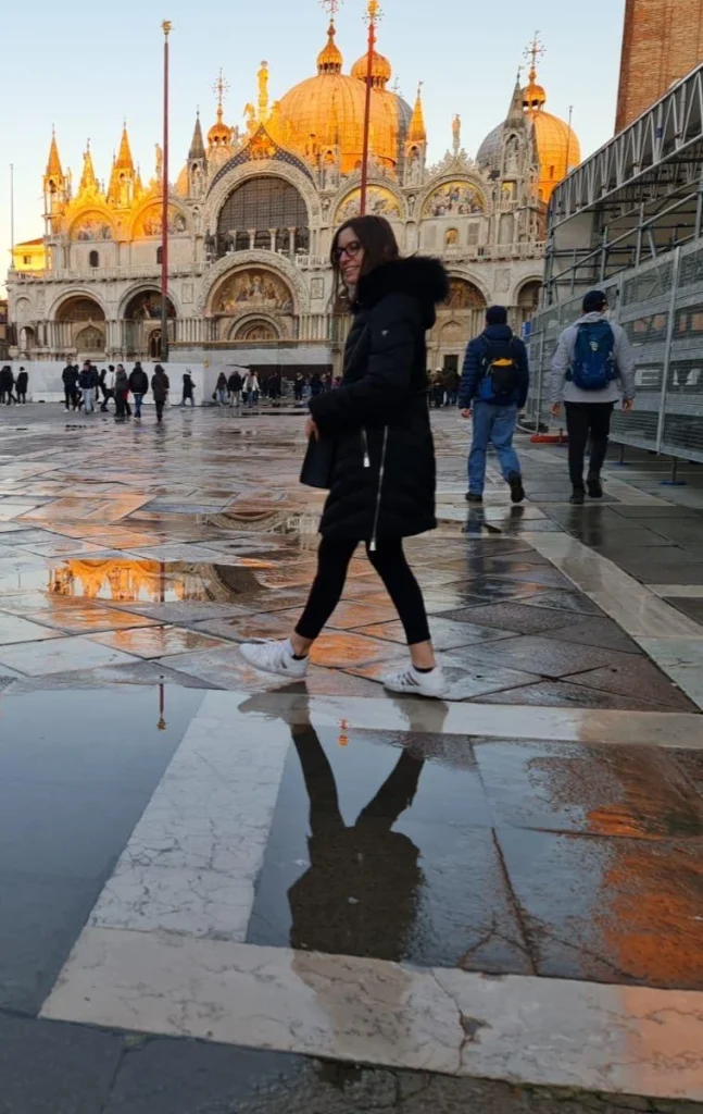 Un momento di riflessione a Venezia. Come una pavese trapiantata in Veneto, anche io trovo il mio riflesso nelle acque di questa città magica, proprio come il riflesso di questa ragazza nella pozzanghera. La maestosa Basilica di San Marco sullo sfondo mi ricorda la bellezza e la ricchezza culturale che mi circondano, sia a Pavia che in Veneto. Un equilibrio perfetto tra le mie radici e la mia nuova casa, proprio come l'armonia tra la figura in primo piano e la magnificenza della basilica.
