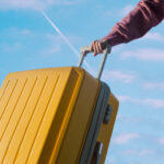 Valigia gialla pronta per un viaggio, con la mano che la tiene saldamente sotto un cielo sereno. Rappresenta la connettività globale e un'esperienza di viaggio senza preoccupazioni per la connessione.