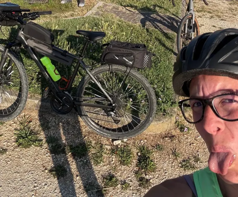 Un primo piano in un selfie di una donna con casco nero e occhiali, che fa la linguaccia in modo scherzoso. Dietro di lei è parcheggiata una bicicletta scura, probabilmente una mountain bike o ibrida, con una borsa da telaio, una borraccia verde neon e una borsa da portapacchi posteriore, appoggiata su un prato a bordo strada.