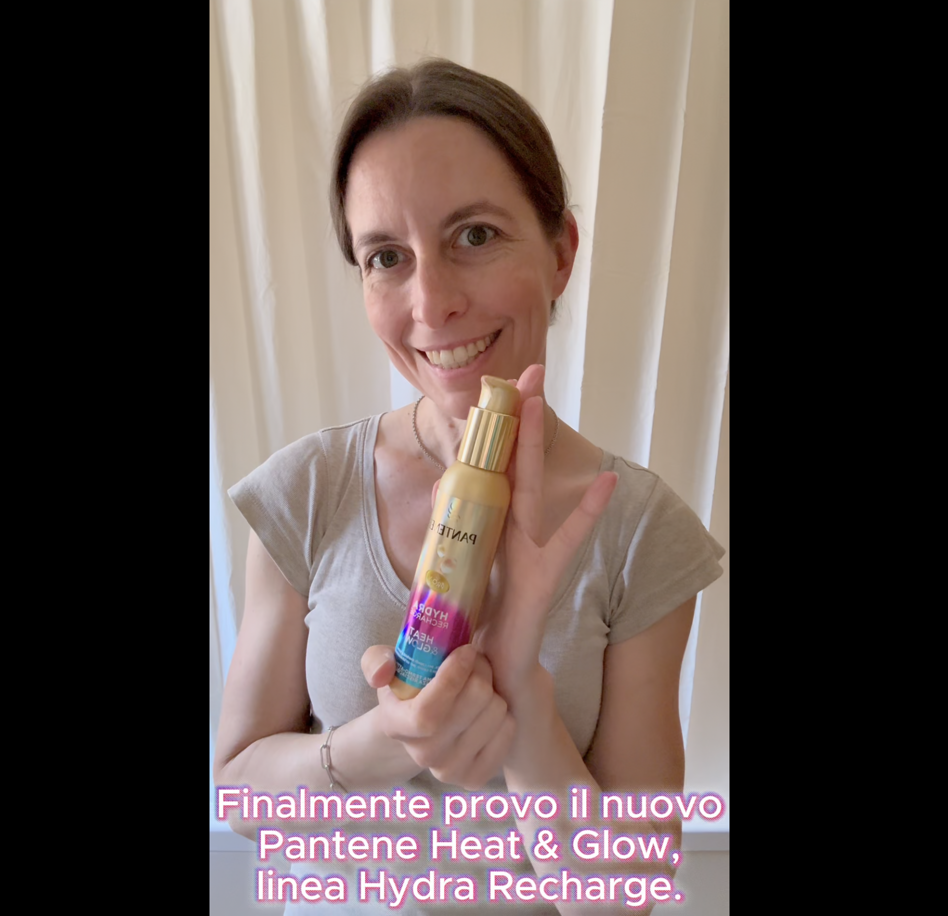 Una donna sorridente tiene in mano il flacone dorato del termoprotettore Pantene Heat & Glow della linea Hydra Recharge. In basso è visibile la scritta con i sottotitoli del video.